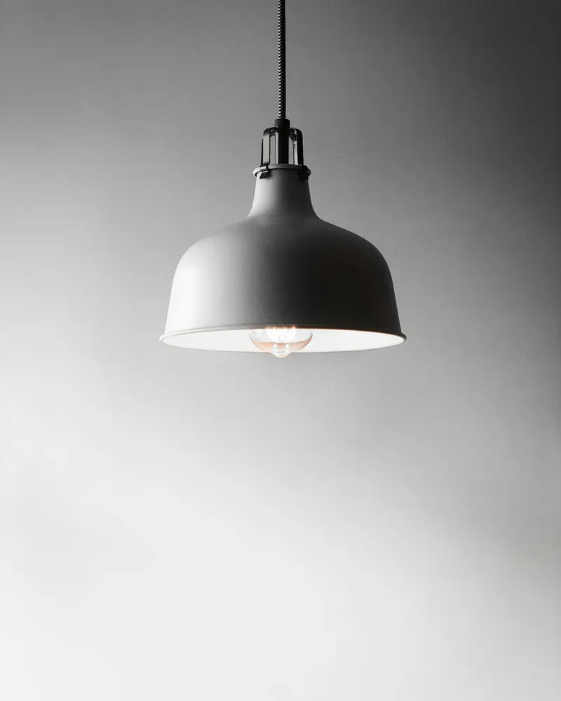 A single matte grey bell-shaped pendant lamp hanging from a black cord against a plain grey wall, bulb visible underneath.