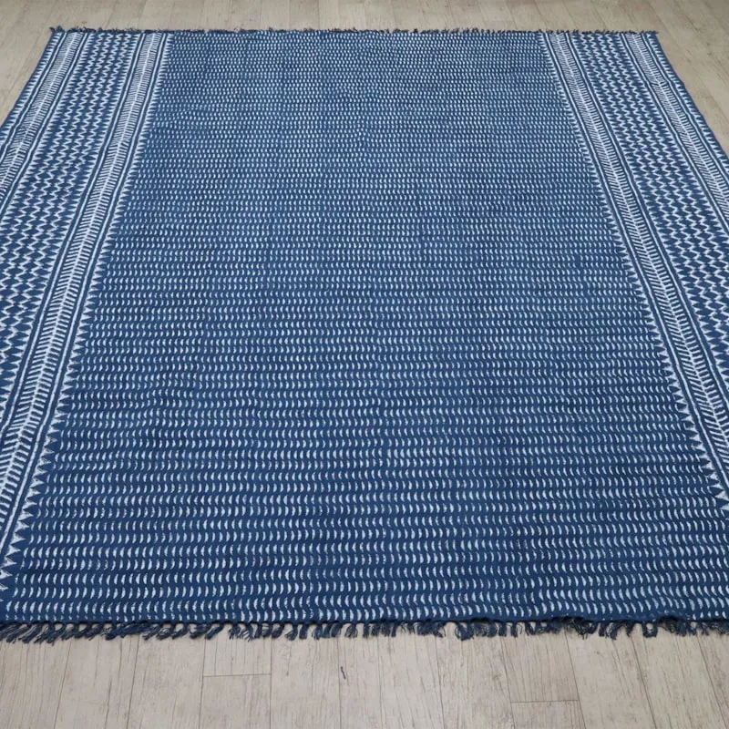 A rectangular flatwoven rug in deep indigo blue with cream chevron-patterned borders, laid flat on a light wood floor.