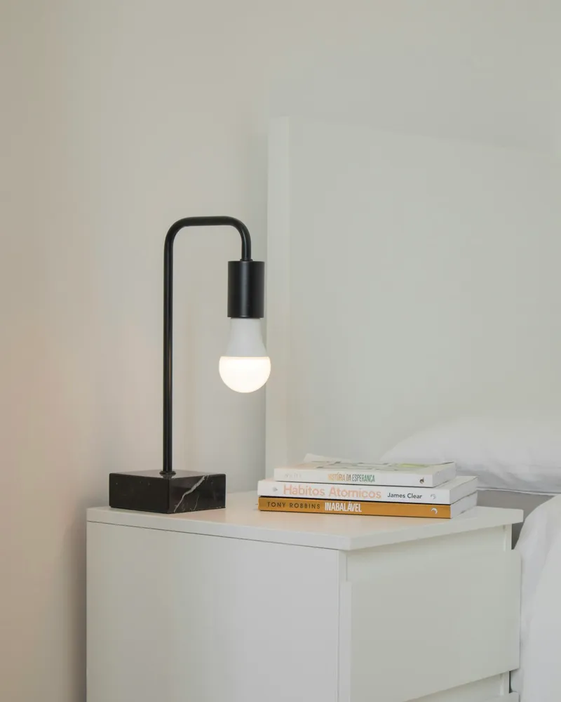 A black right-angled portable lamp with an exposed frosted bulb and a square marble base, on a white nightstand next to a stack of books.
