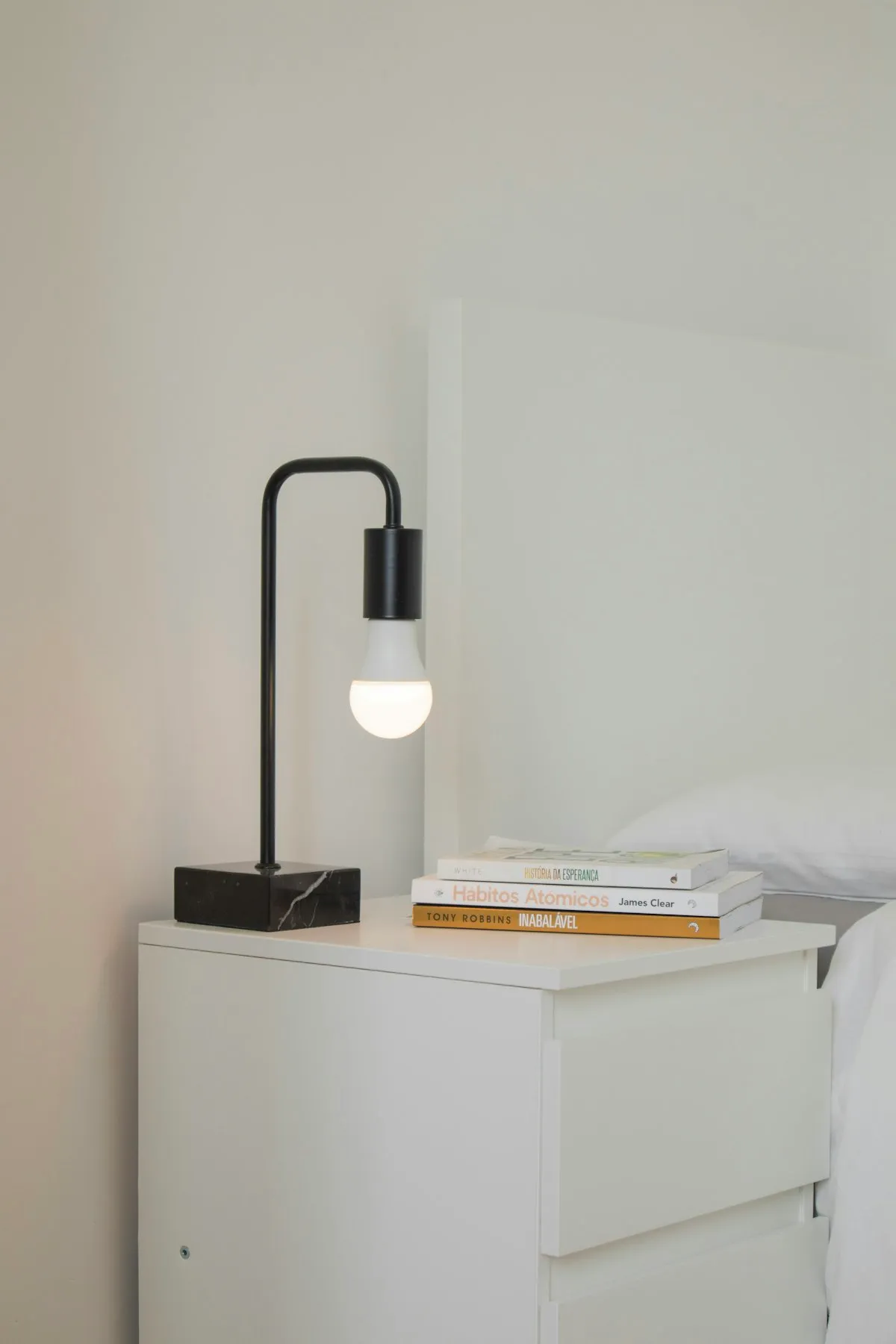A black right-angled portable lamp with an exposed frosted bulb and a square marble base, on a white nightstand next to a stack of books.
