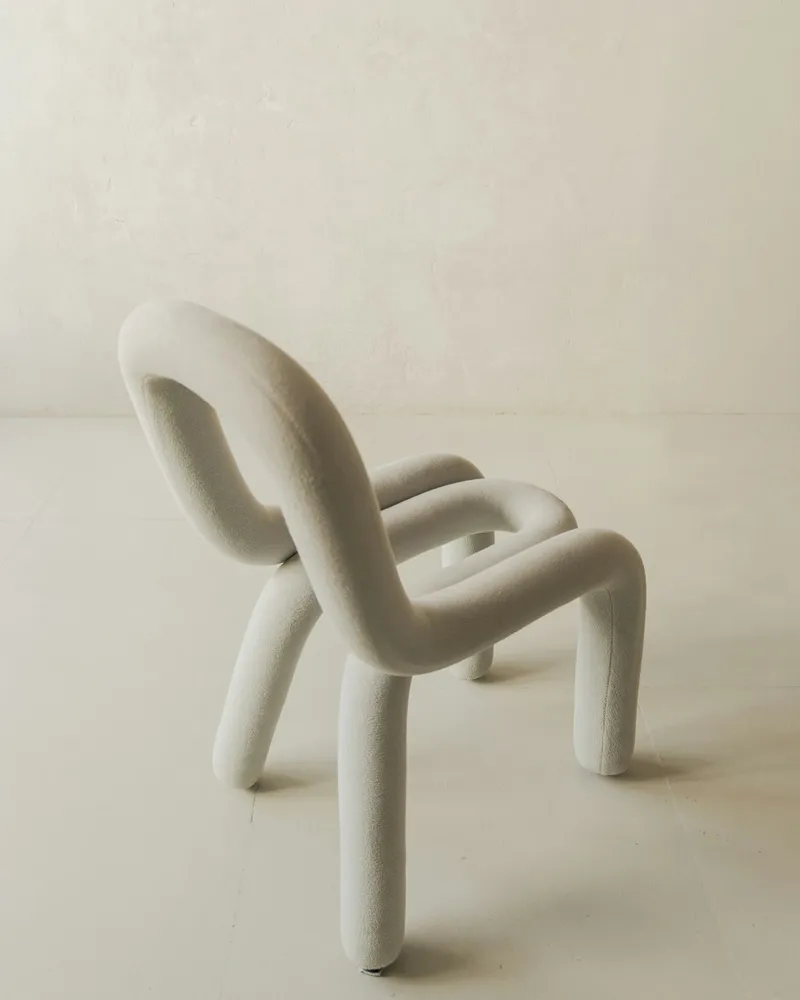 Sculptural bouclé lounge chair formed from one continuous looping tube shape that folds into a seat, backrest, arms, and four legs, on a plain light studio floor and wall.