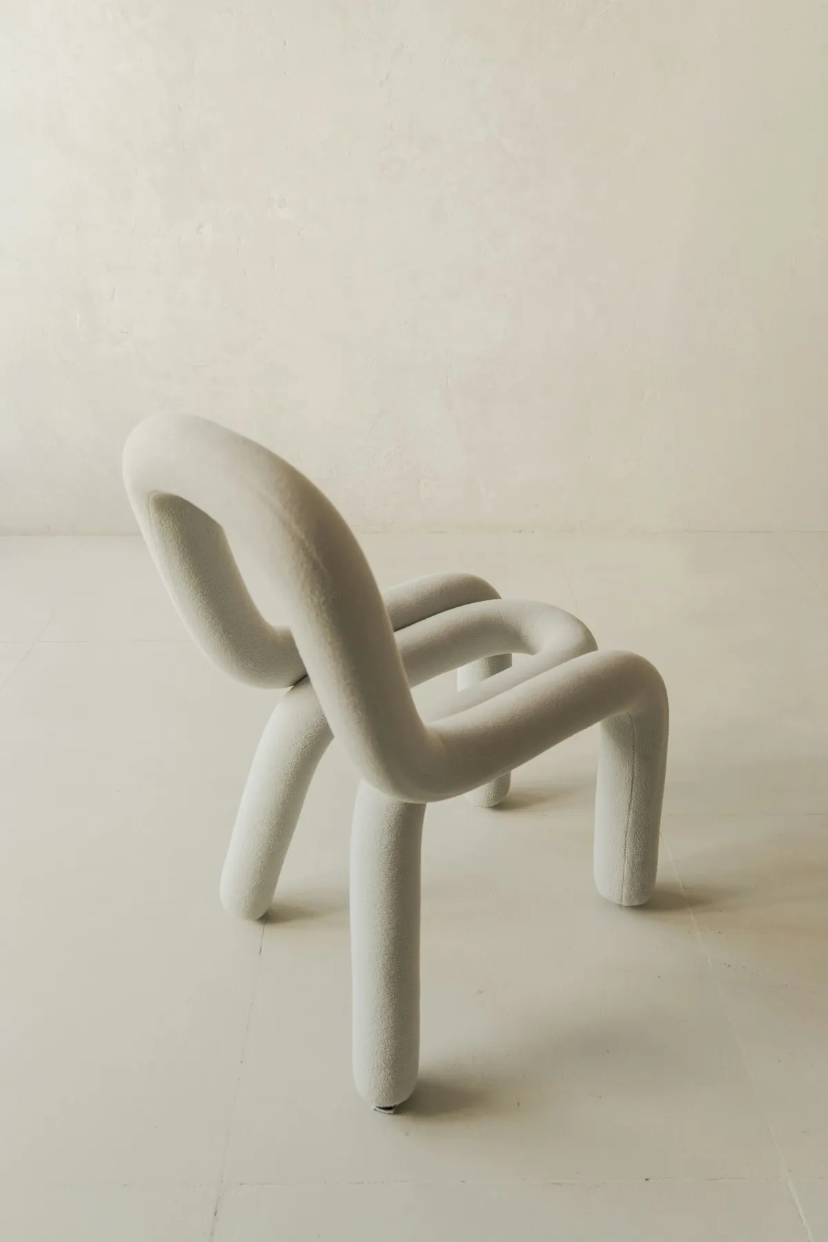 Sculptural bouclé lounge chair formed from one continuous looping tube shape that folds into a seat, backrest, arms, and four legs, on a plain light studio floor and wall.