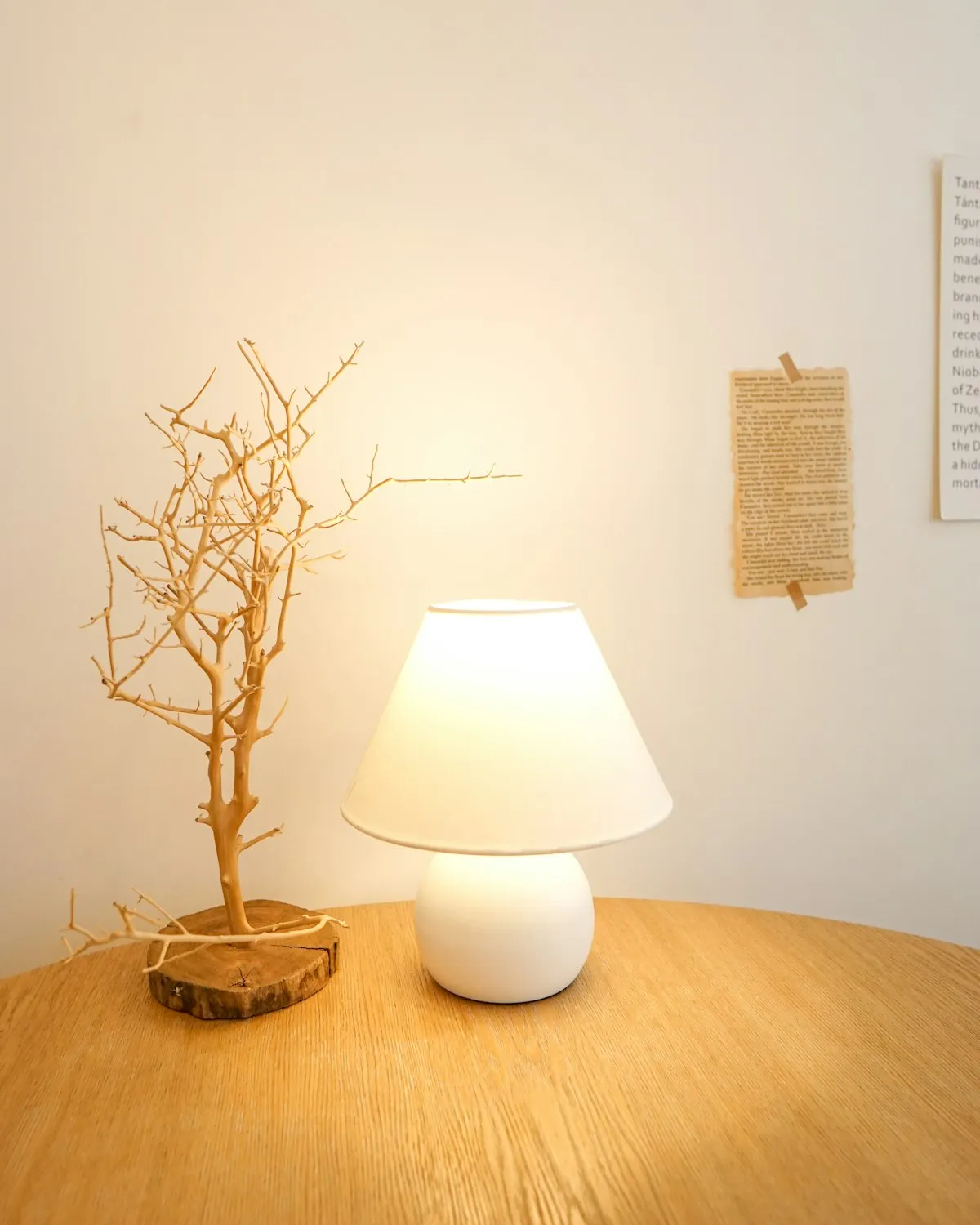 A round ceramic table lamp with a smooth pebble-shaped white base and a tapered fabric shade, glowing on a wooden table next to a dried branch ornament.