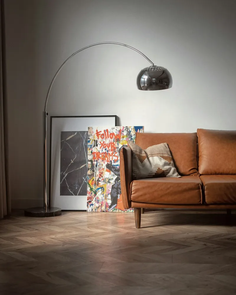 A chrome arc floor lamp with a domed metal shade, curving over a leather sofa in front of a marble-based standing lamp and framed artwork.