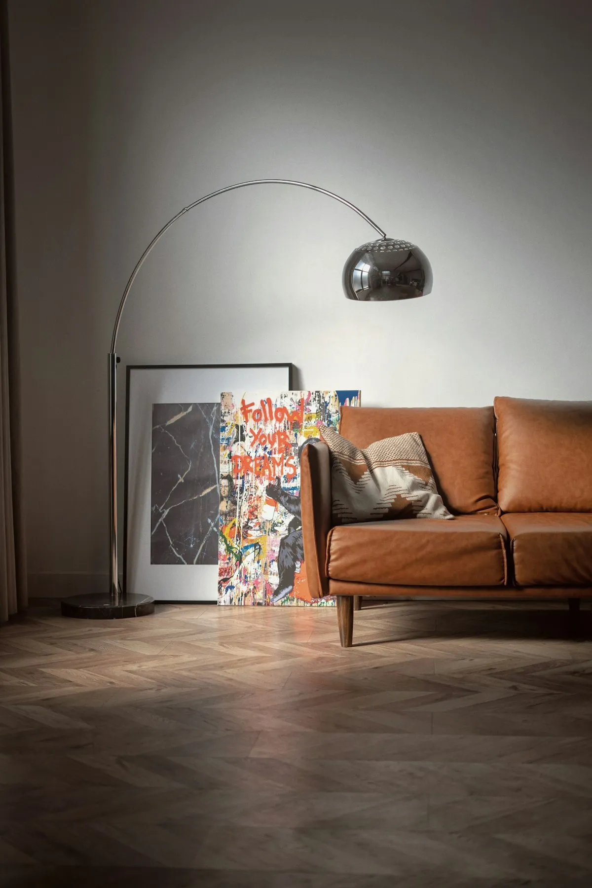 A chrome arc floor lamp with a domed metal shade, curving over a leather sofa in front of a marble-based standing lamp and framed artwork.