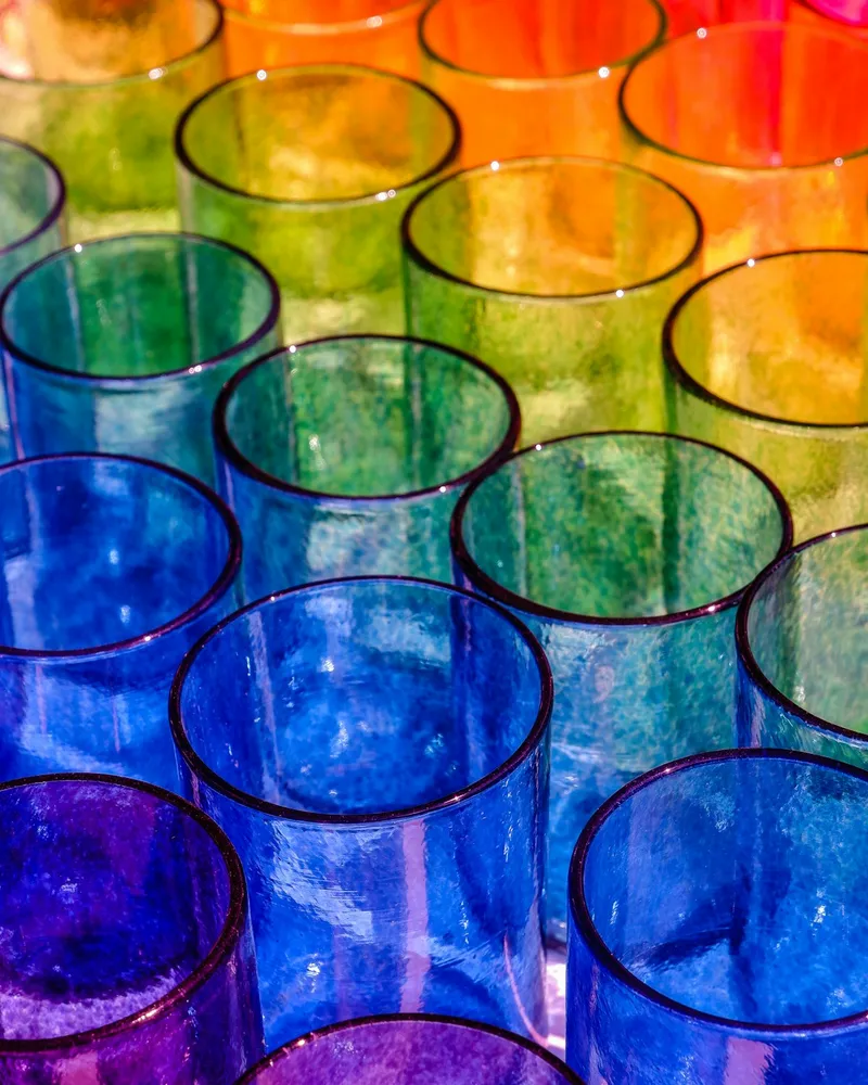 Overhead view of an array of hand-blown drinking glasses in red, orange, green, blue, and purple tinted glass, arranged in rows on a white surface with dramatic natural light casting long shadows.