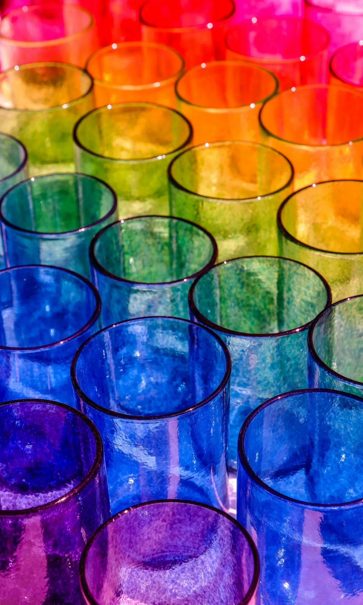 Overhead view of an array of hand-blown drinking glasses in red, orange, green, blue, and purple tinted glass, arranged in rows on a white surface with dramatic natural light casting long shadows.