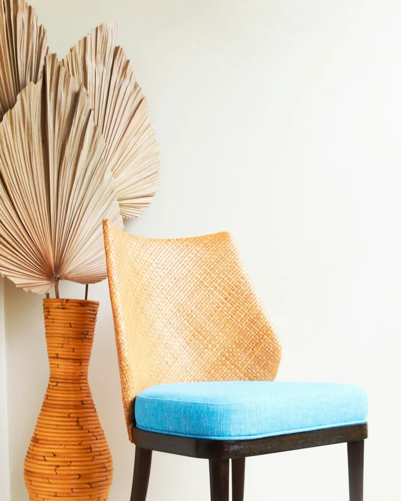 Dining chair with a tall tapered woven cane backrest, a sky-blue upholstered seat cushion, and dark stained wooden legs, against a plain wall with a woven vase and dried palm fronds beside it.