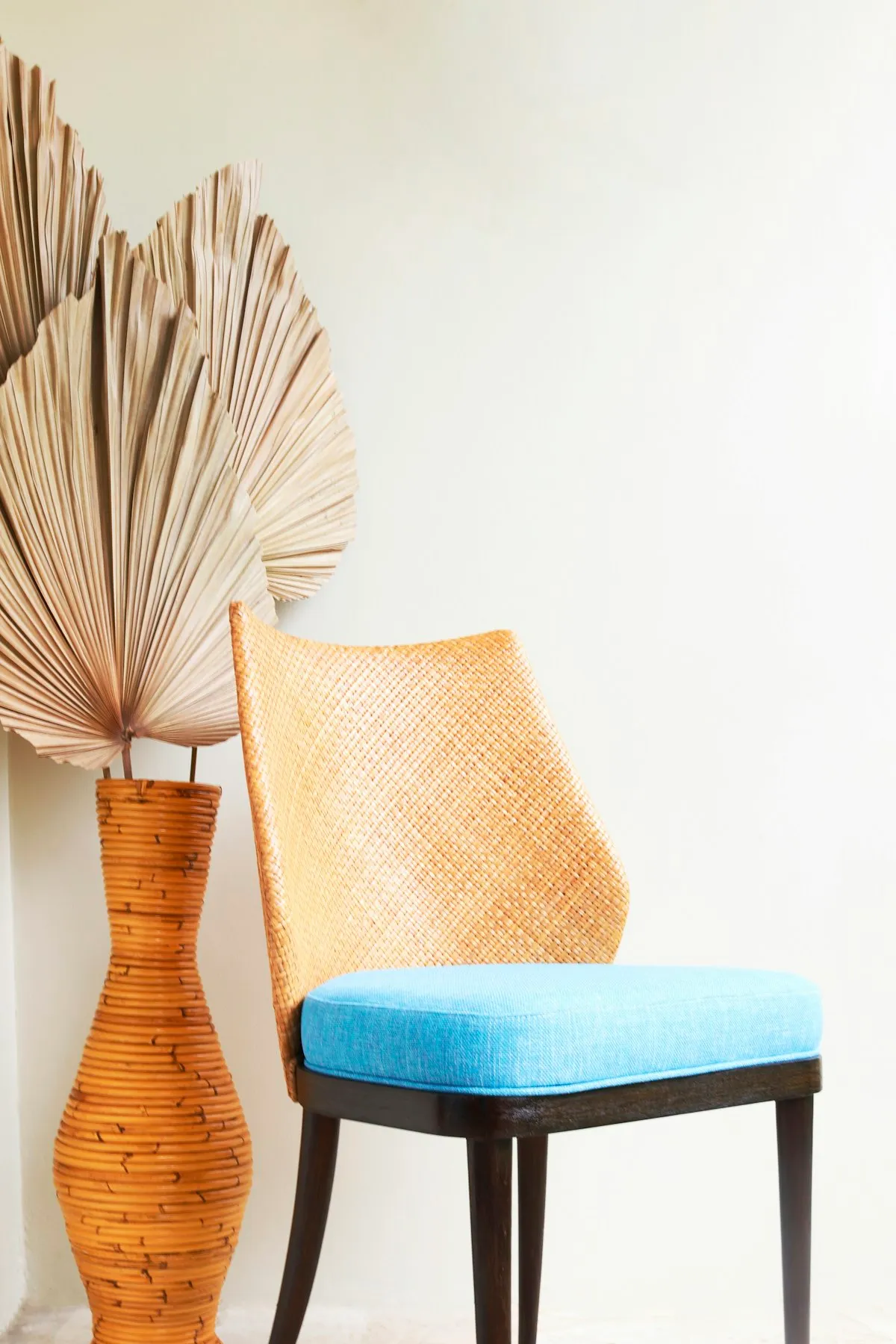 Dining chair with a tall tapered woven cane backrest, a sky-blue upholstered seat cushion, and dark stained wooden legs, against a plain wall with a woven vase and dried palm fronds beside it.