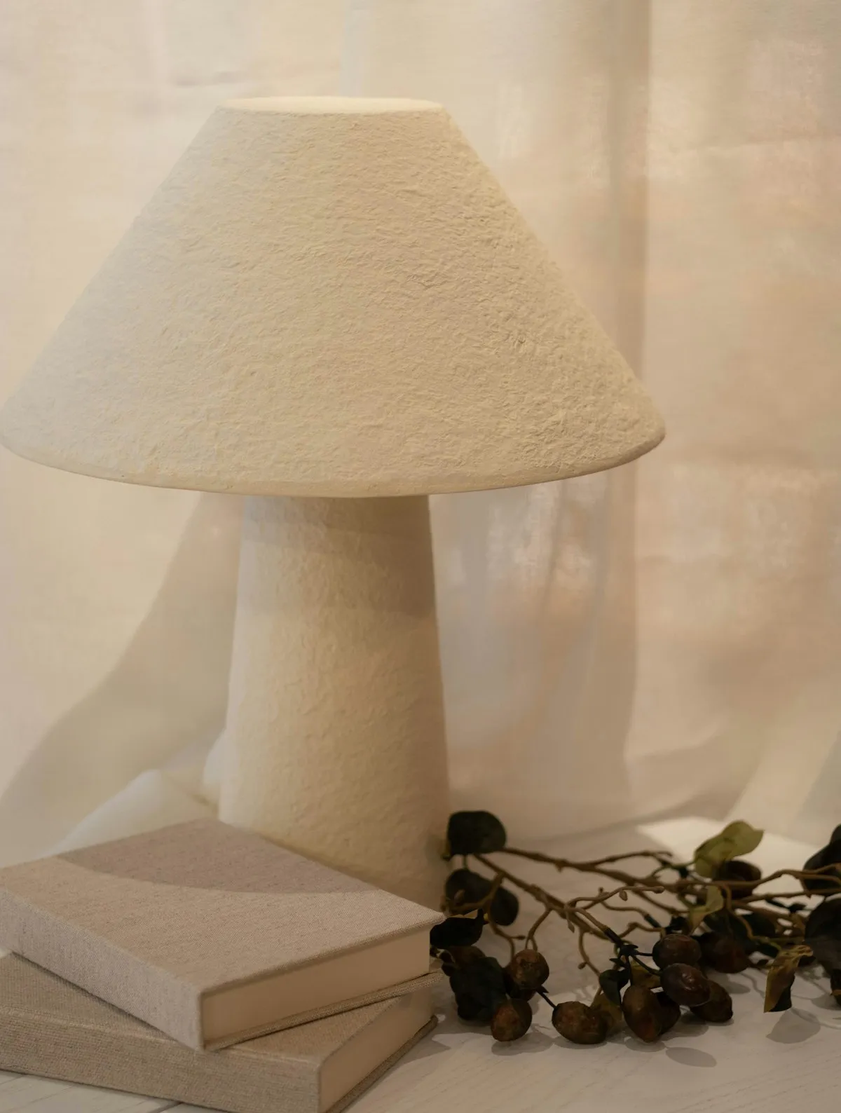 A white, rough-textured plaster table lamp with a conical shade and tapered base, standing in front of a sheer linen curtain.
