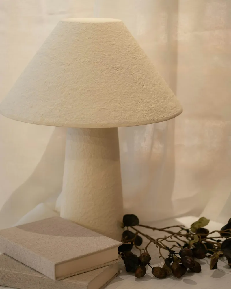 A white, rough-textured plaster table lamp with a conical shade and tapered base, standing in front of a sheer linen curtain.