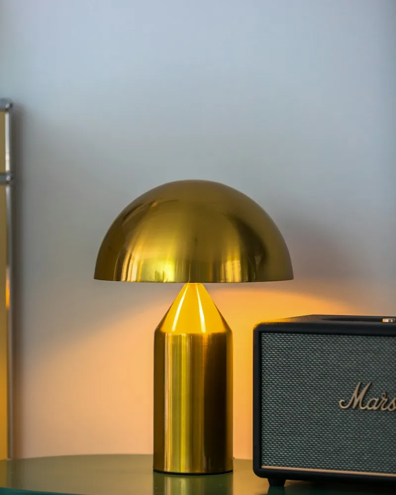 A brass mushroom-dome table lamp with a cylindrical base, glowing warmly on a green sideboard next to a black speaker.
