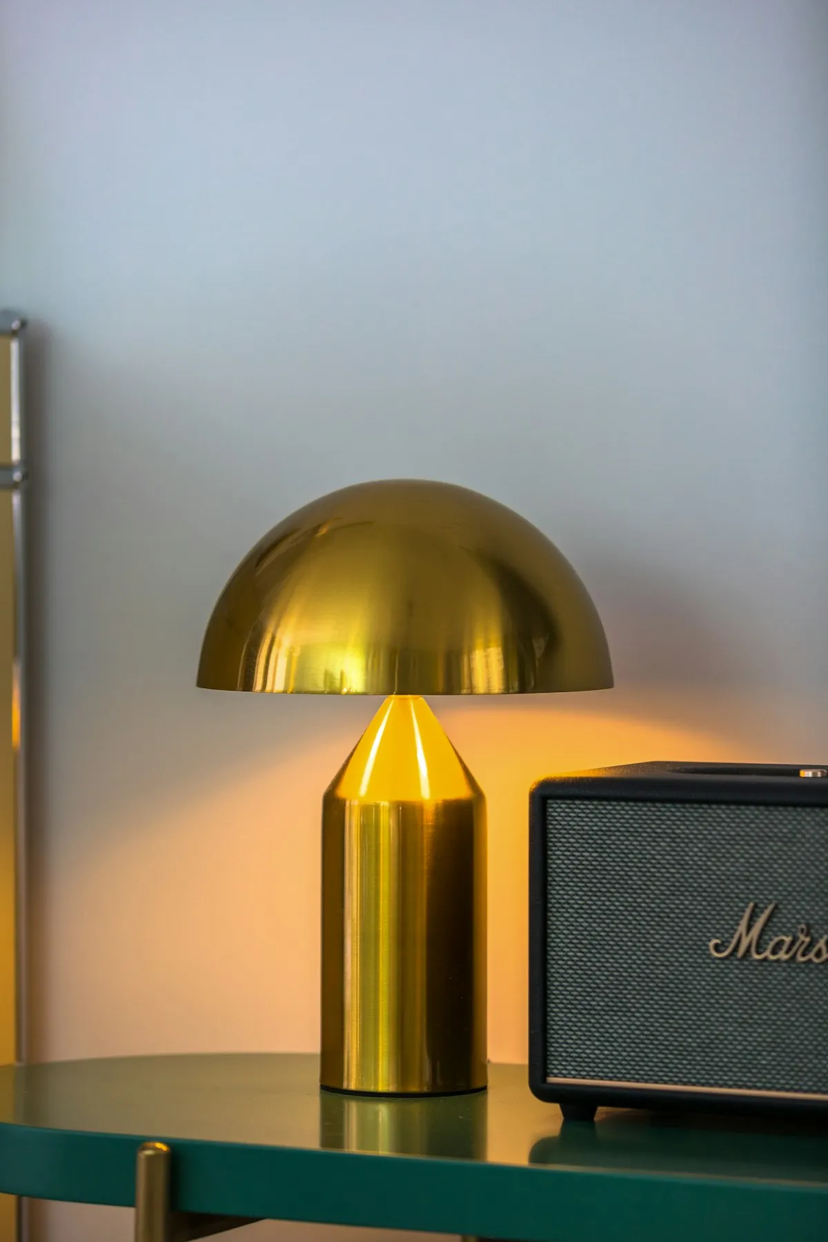 A brass mushroom-dome table lamp with a cylindrical base, glowing warmly on a green sideboard next to a black speaker.