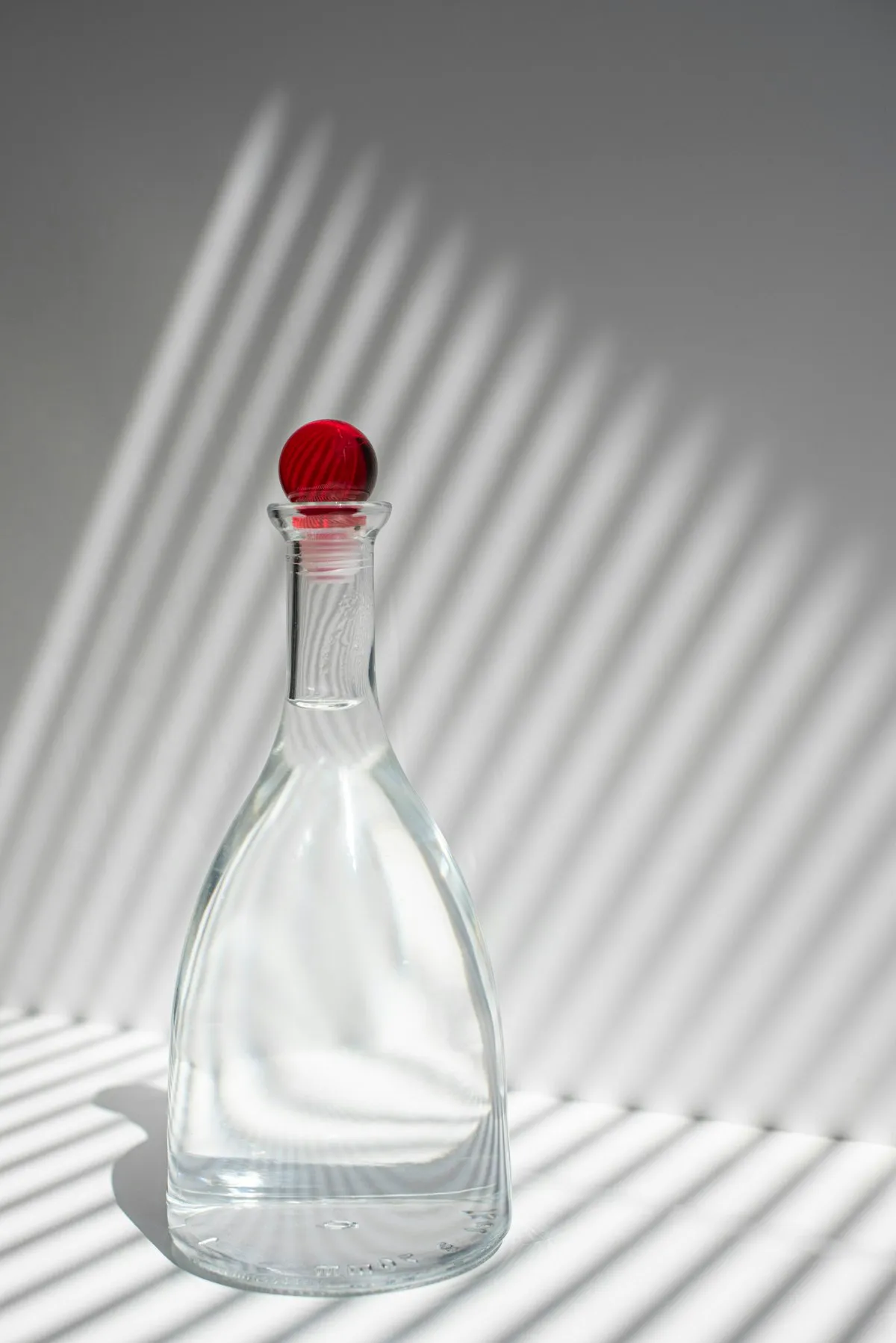 Clear glass carafe with a tapered faceted body and a round red glass stopper, photographed against a plain grey backdrop with strong striped shadow lines.