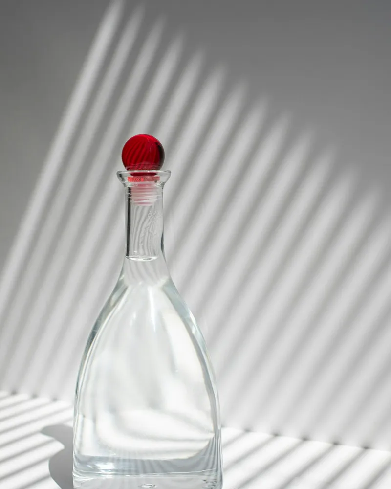 Clear glass carafe with a tapered faceted body and a round red glass stopper, photographed against a plain grey backdrop with strong striped shadow lines.