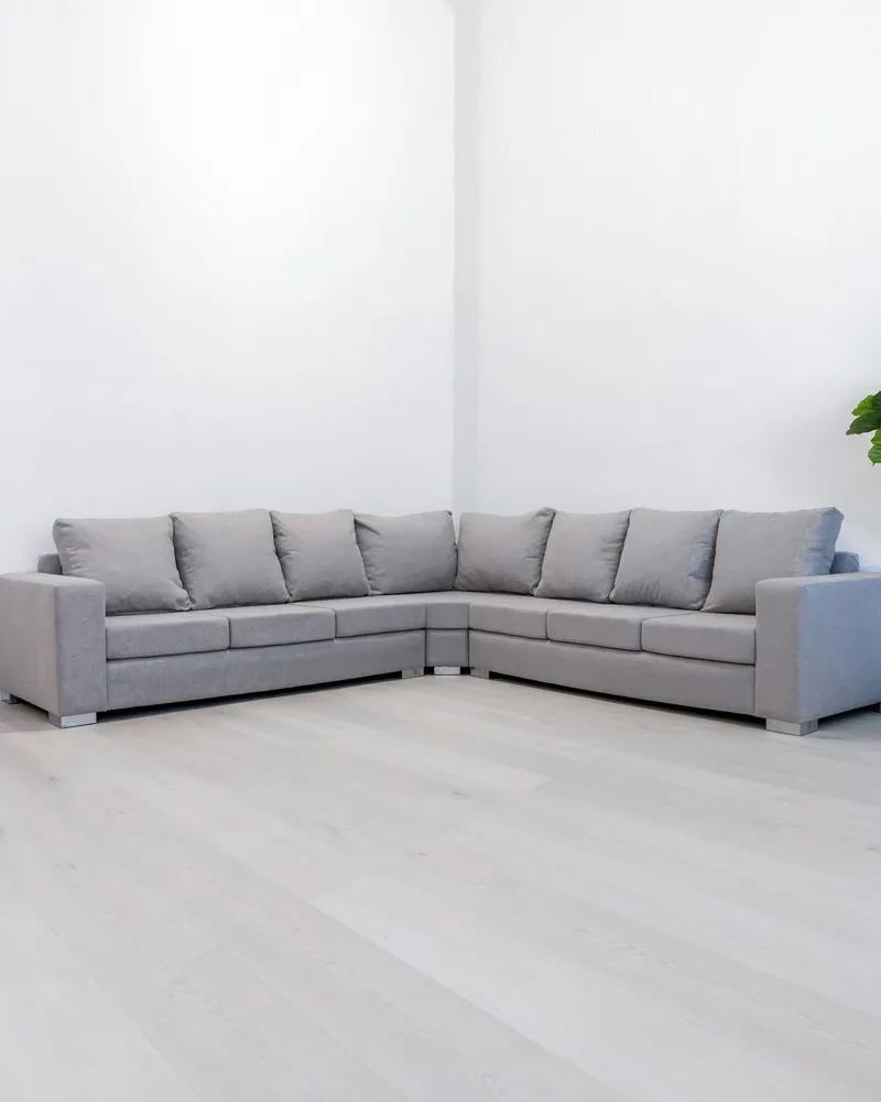 Grey L-shaped modular sectional sofa with boxy square arms and chrome feet, arranged in the corner of a bright white minimal room with light wood flooring.