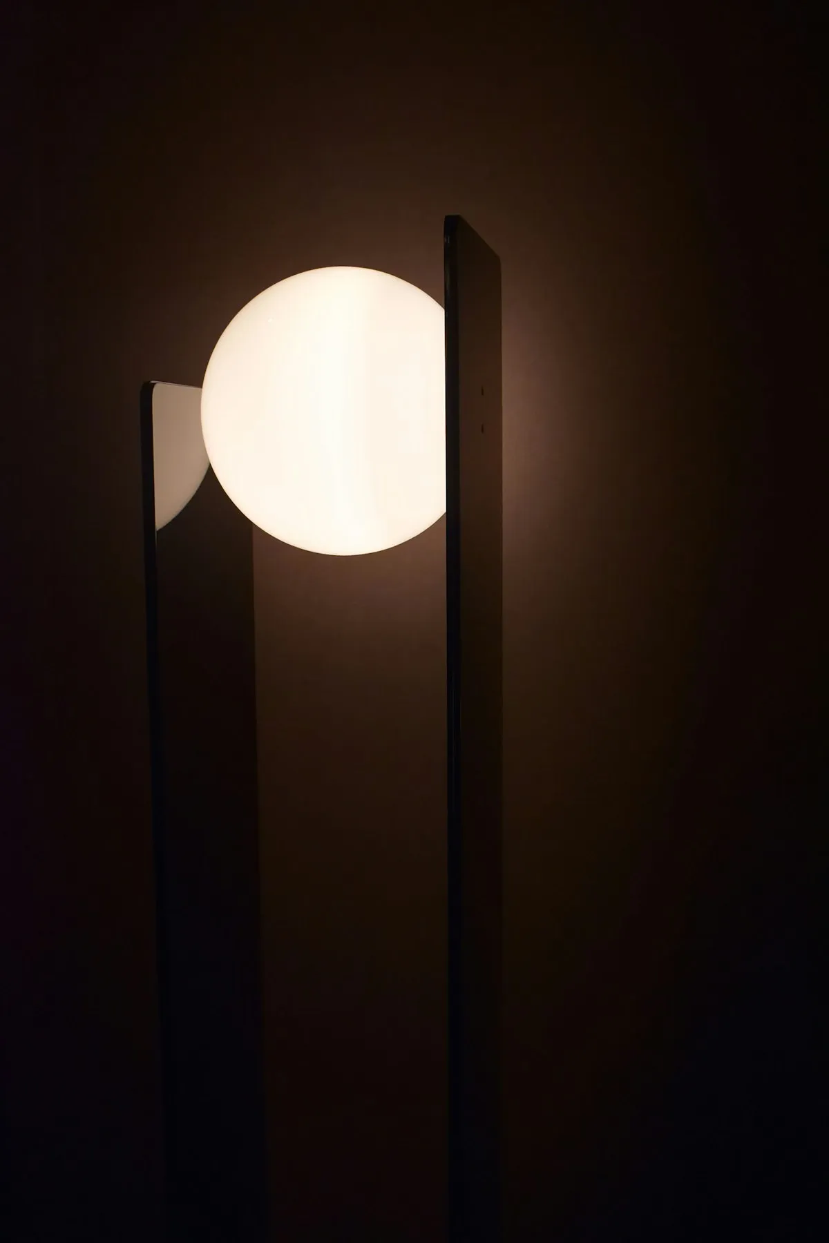 A tall, dark slab-shaped floor lamp with a single glowing white glass sphere set into its side, standing in a dim room.