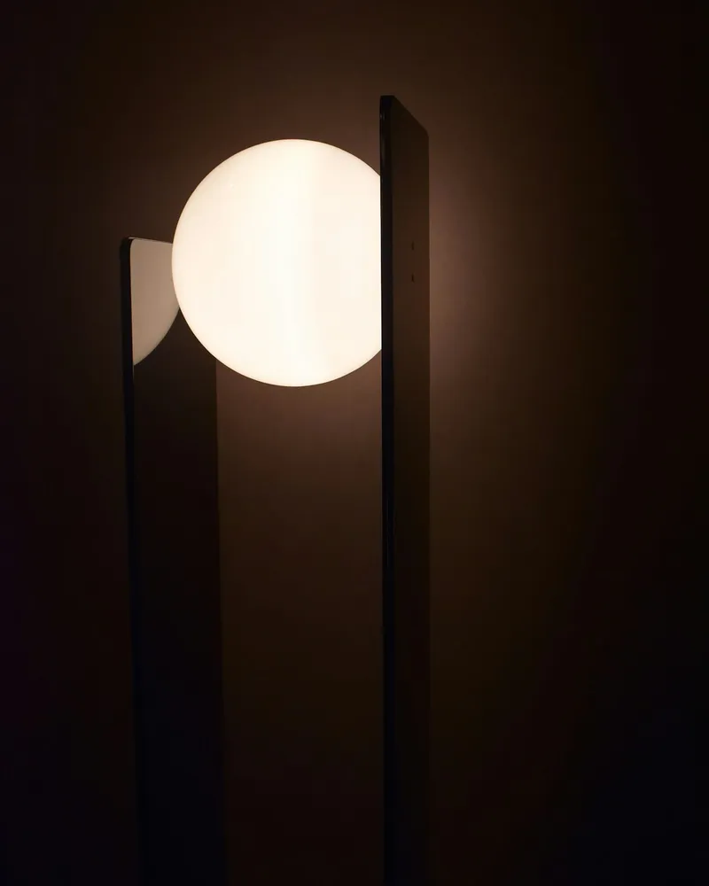 A tall, dark slab-shaped floor lamp with a single glowing white glass sphere set into its side, standing in a dim room.