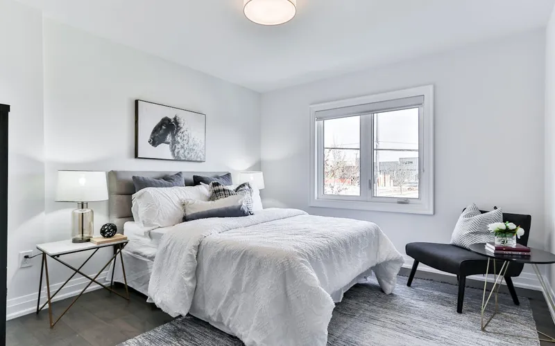 Bright, minimal bedroom with white bedding, a grey upholstered headboard, and a reading chair by the window