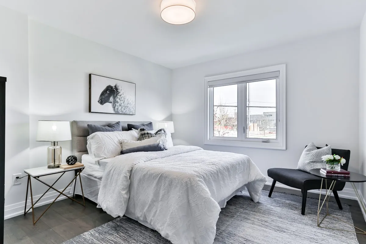 Bright, minimal bedroom with white bedding, a grey upholstered headboard, and a reading chair by the window