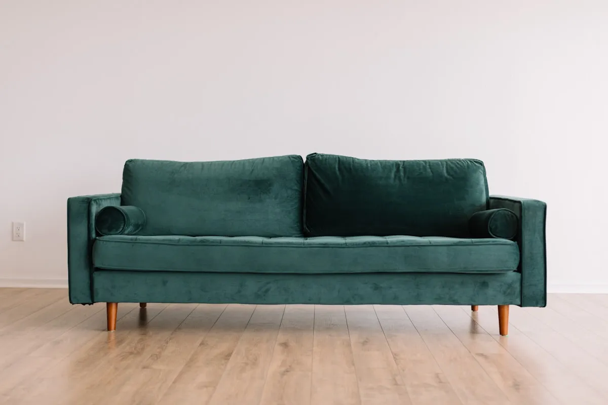 Emerald green velvet two-seat sofa with tufted seat cushion, bolster arm pillows, and tapered wooden legs, against a plain white wall on a light wood floor.