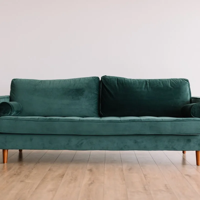 Emerald green velvet two-seat sofa with tufted seat cushion, bolster arm pillows, and tapered wooden legs, against a plain white wall on a light wood floor.