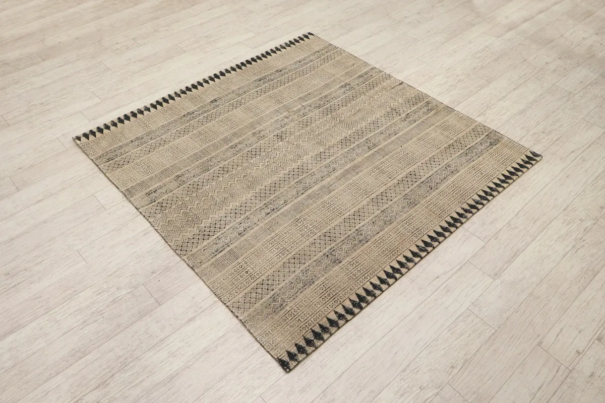 A tan and black flatwoven rug with narrow striped and geometric banding, laid at an angle on a light wood floor.
