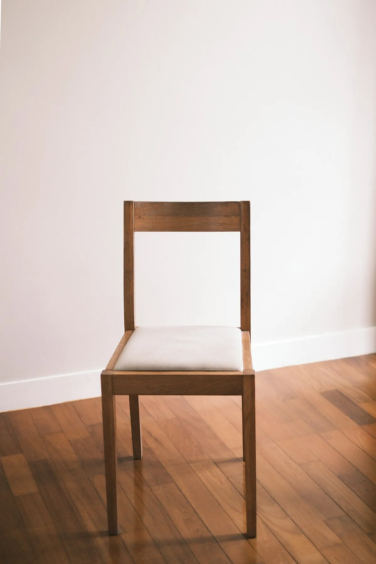Simple oak dining chair with a plain ladder-style backrest and a stitched cream upholstered seat pad, on a wooden floor against a white wall.