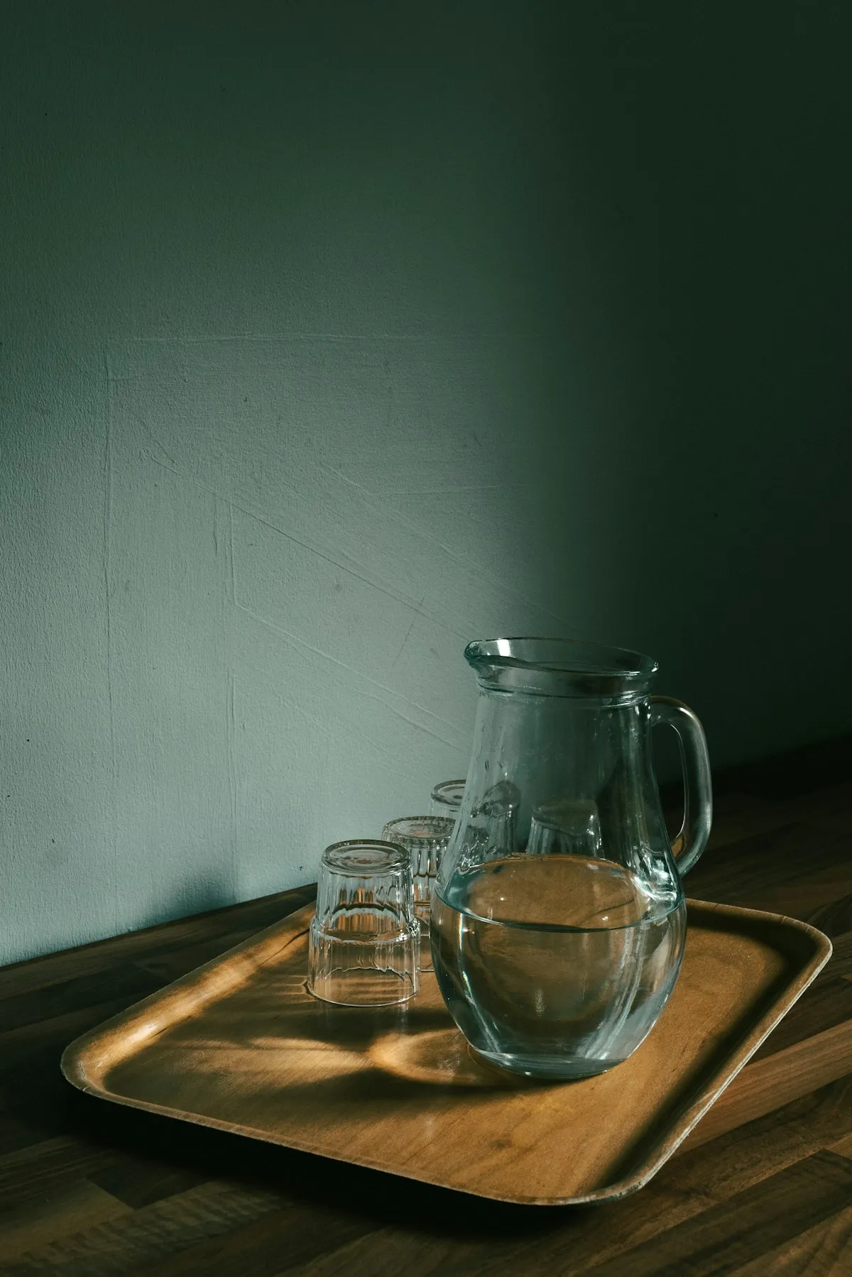 Clear glass pitcher filled with water alongside two small drinking glasses, arranged on a wooden tray against a dark green wall.
