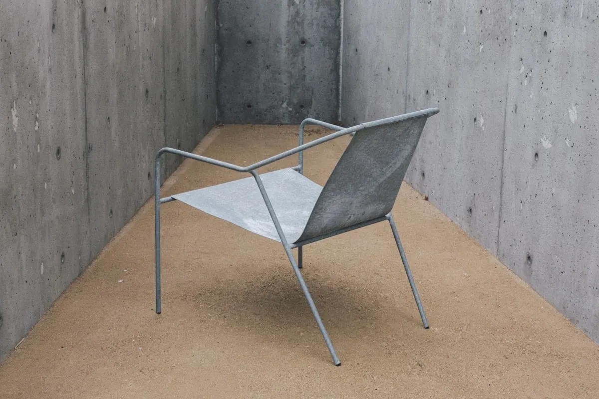 Minimalist grey metal-frame lounge chair with a flat folded sheet-metal seat and backrest, photographed in a bare concrete alcove.