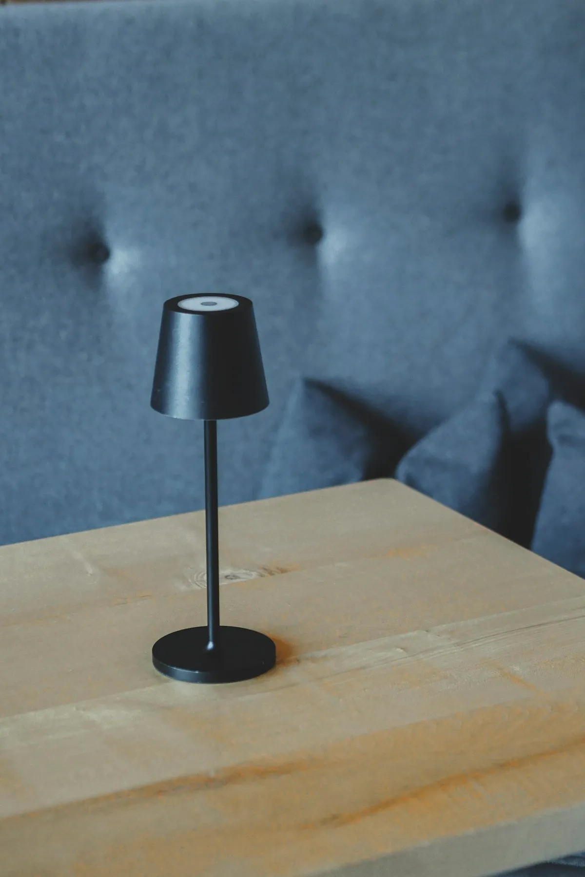 A slim black cordless table lamp with a tapered drum shade, standing on a wooden table against a tufted grey headboard.