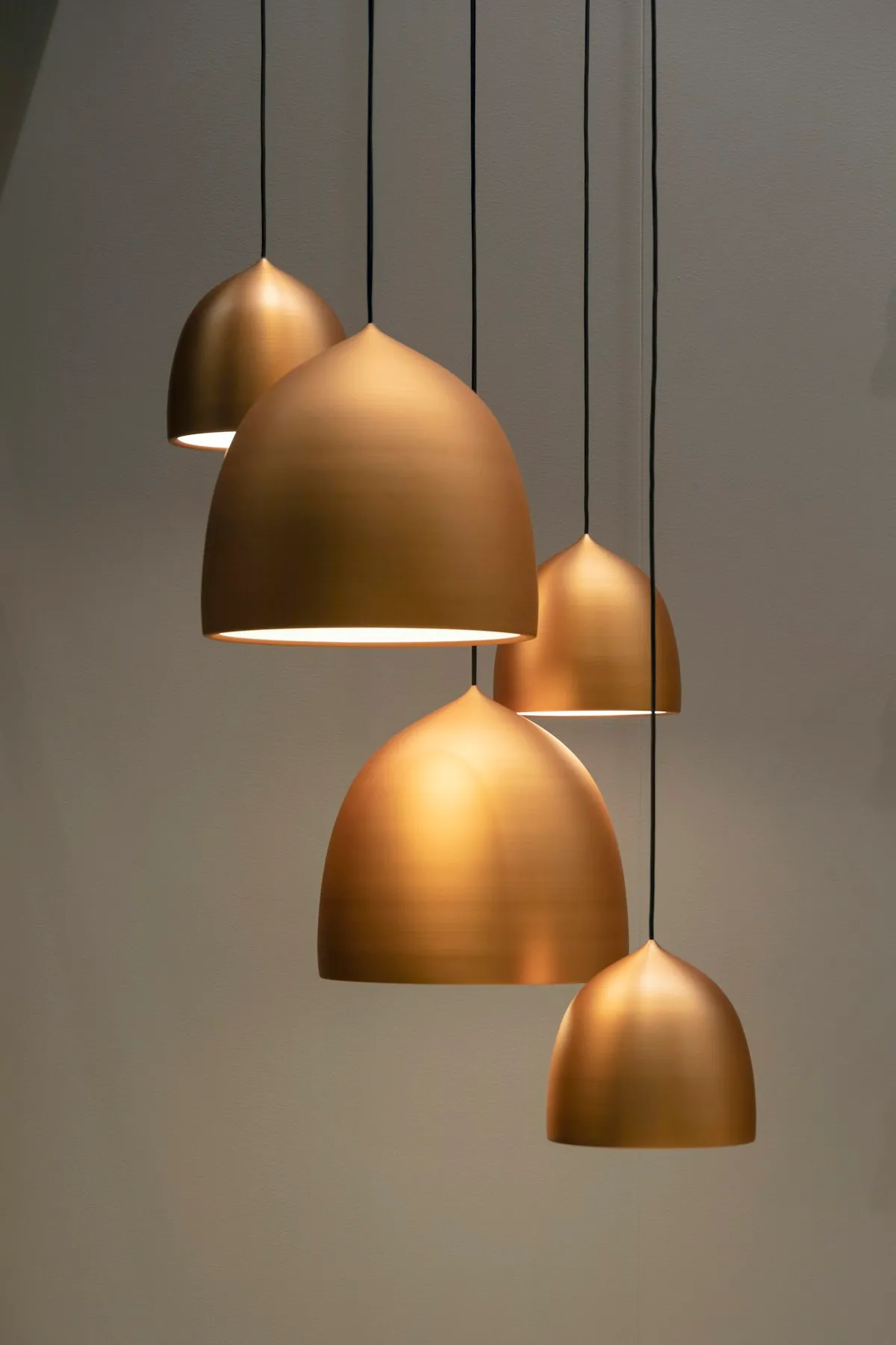Five brass dome pendant lights of varying sizes hanging at different heights against a plain grey wall.