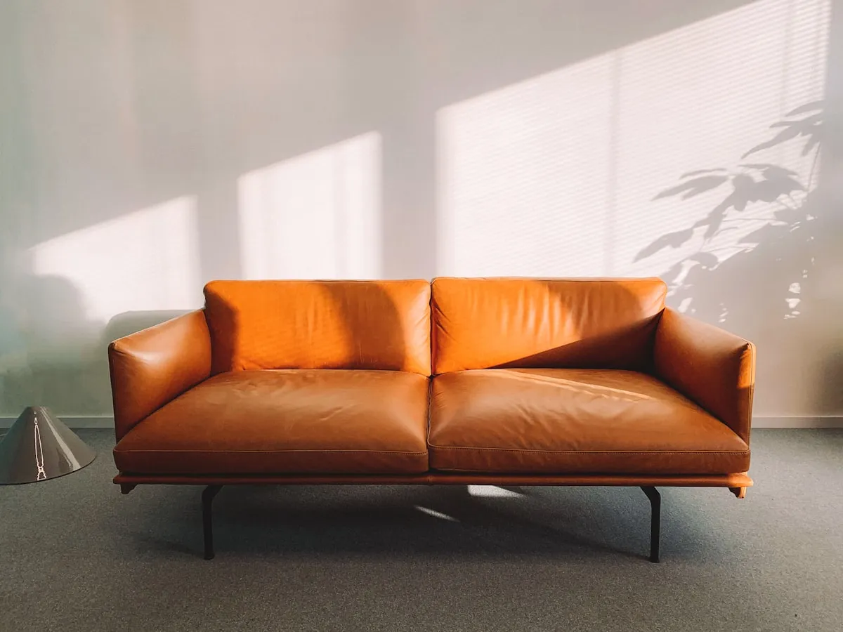 Cognac tan leather two-seat sofa with slim black metal legs, photographed in raking natural light against a plain wall with a plant shadow.