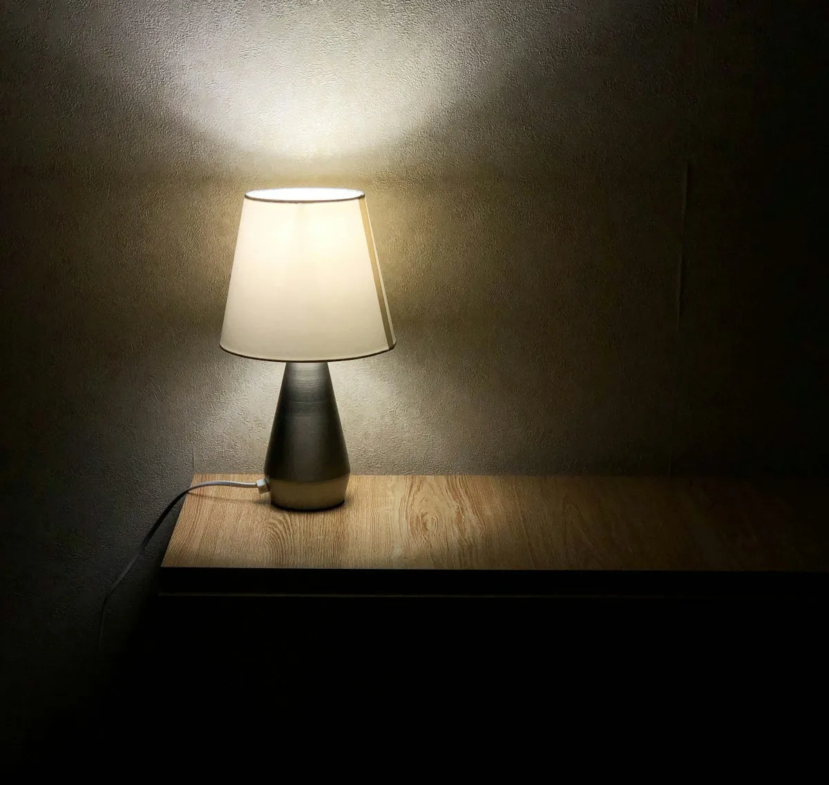 A metallic conical table lamp with a cream fabric drum shade, glowing on a wooden surface against a plain dark wall.