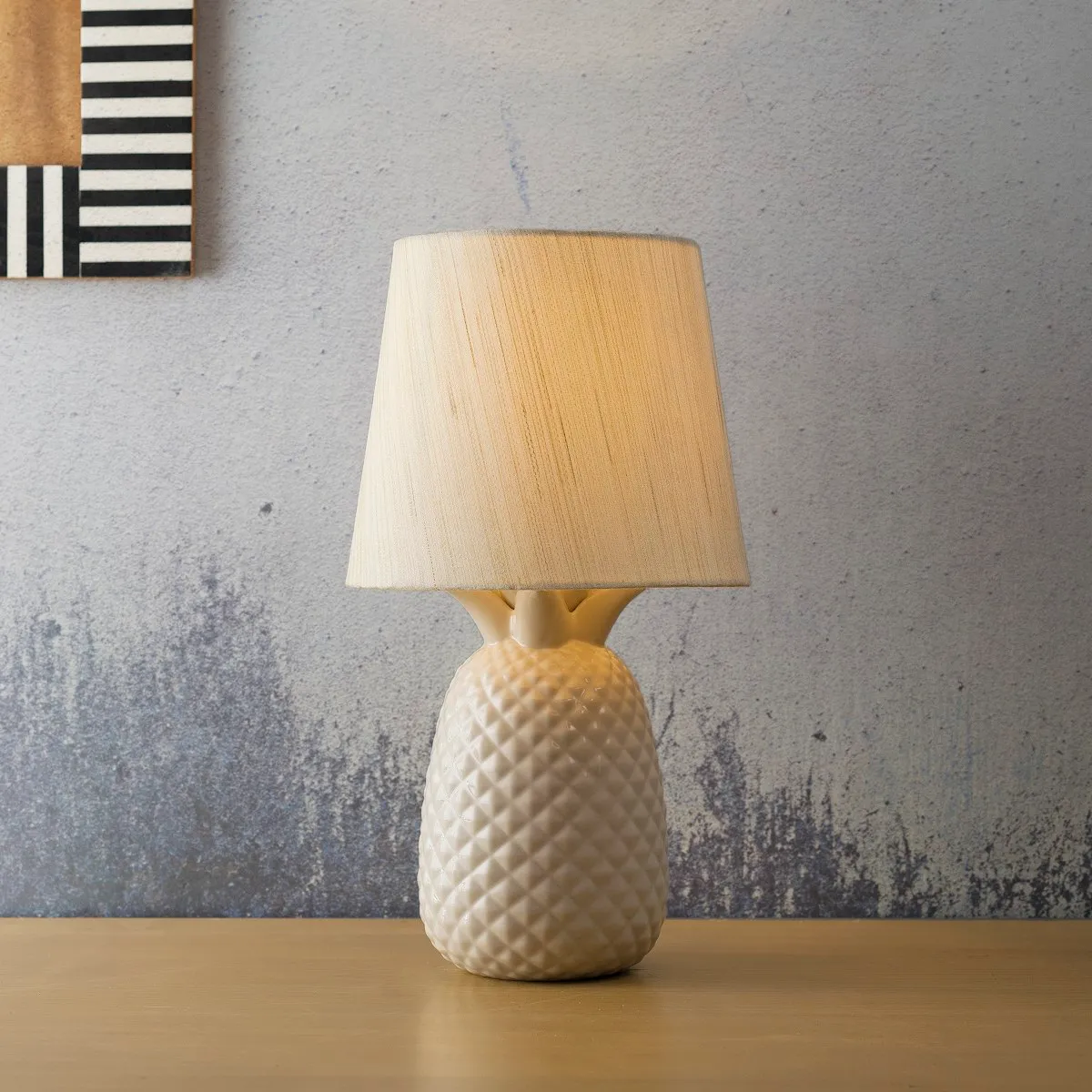 A cream ceramic table lamp with a textured pineapple-patterned base and a tapered fabric shade, against a mottled grey wall.