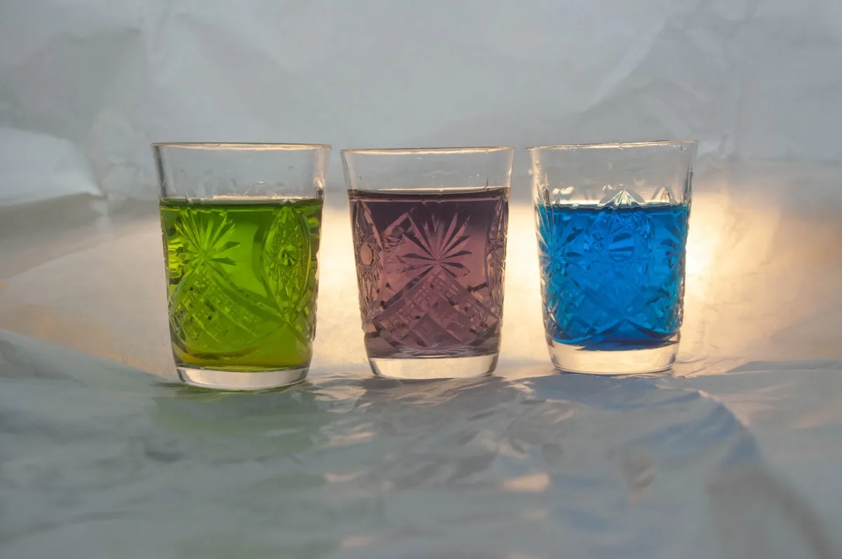 Three faceted drinking glasses filled with green, purple, and blue liquid, lit from behind on a soft white backdrop.