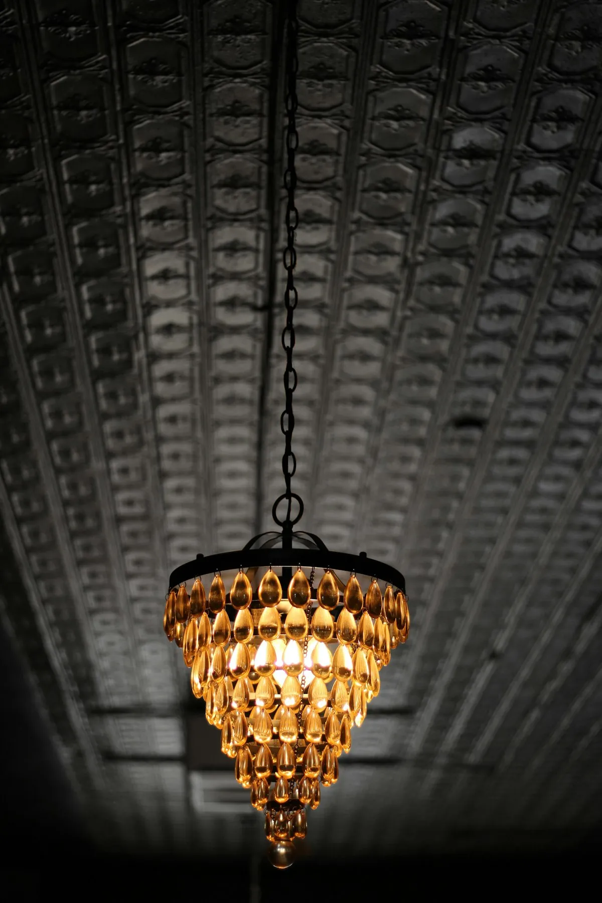 A tiered chandelier made of amber teardrop-shaped glass beads, hanging from a chain against a dark textured ceiling.