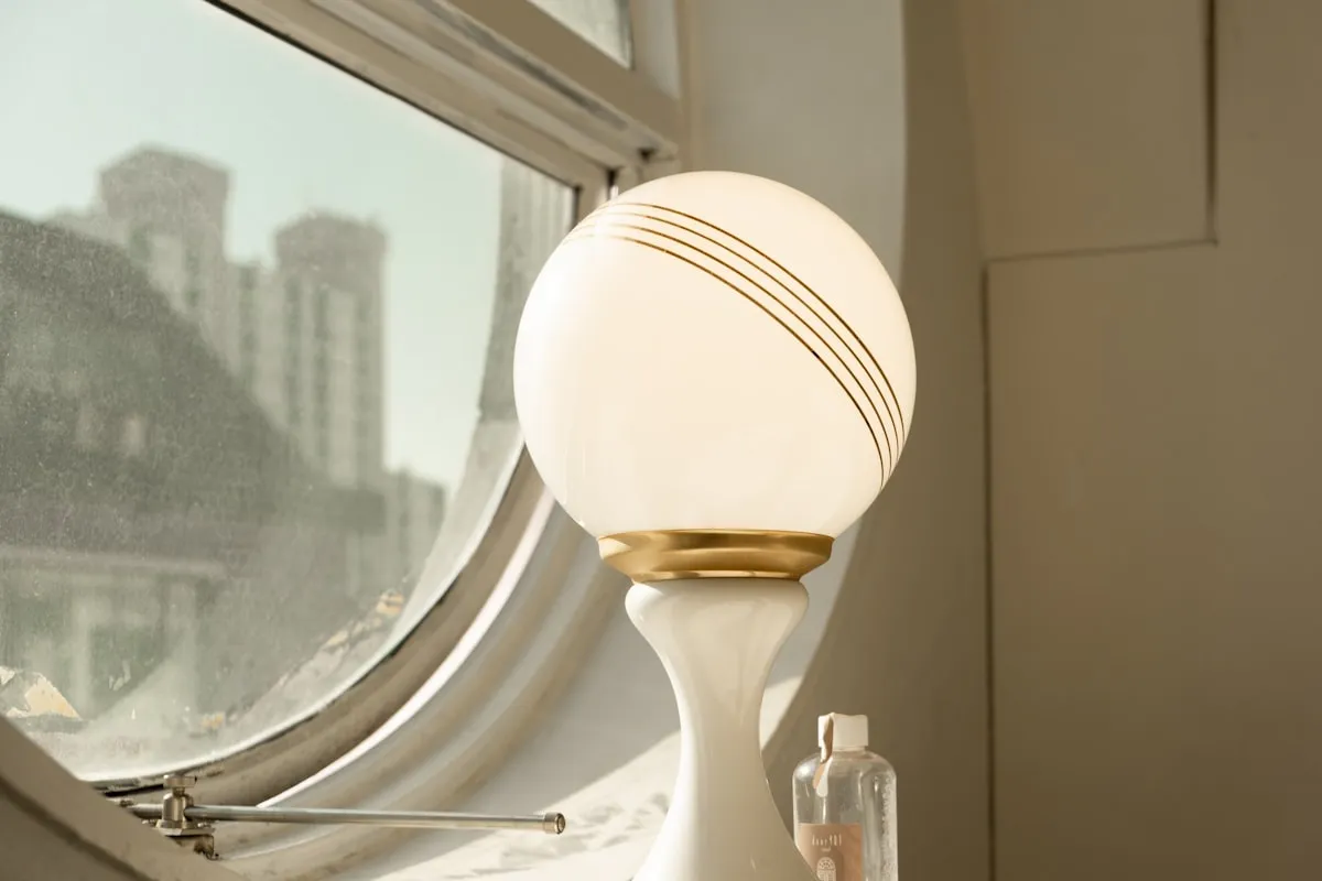 A vintage-style globe table lamp with gold banding on a cream ceramic base, standing beside a window with a city view.