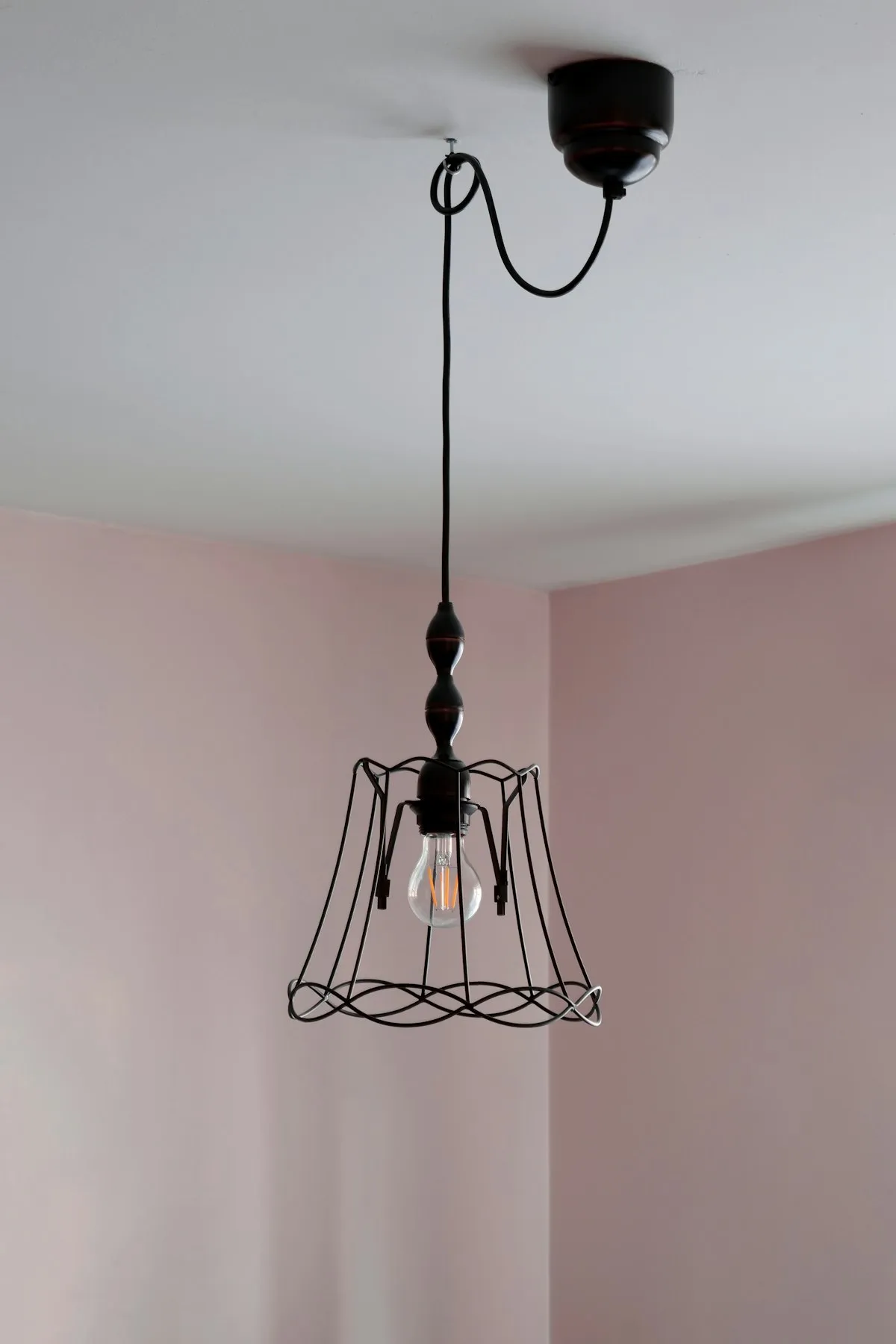 A wire-cage pendant lamp with an exposed bulb, hanging from a dark ceiling fixture against a pale pink wall corner.