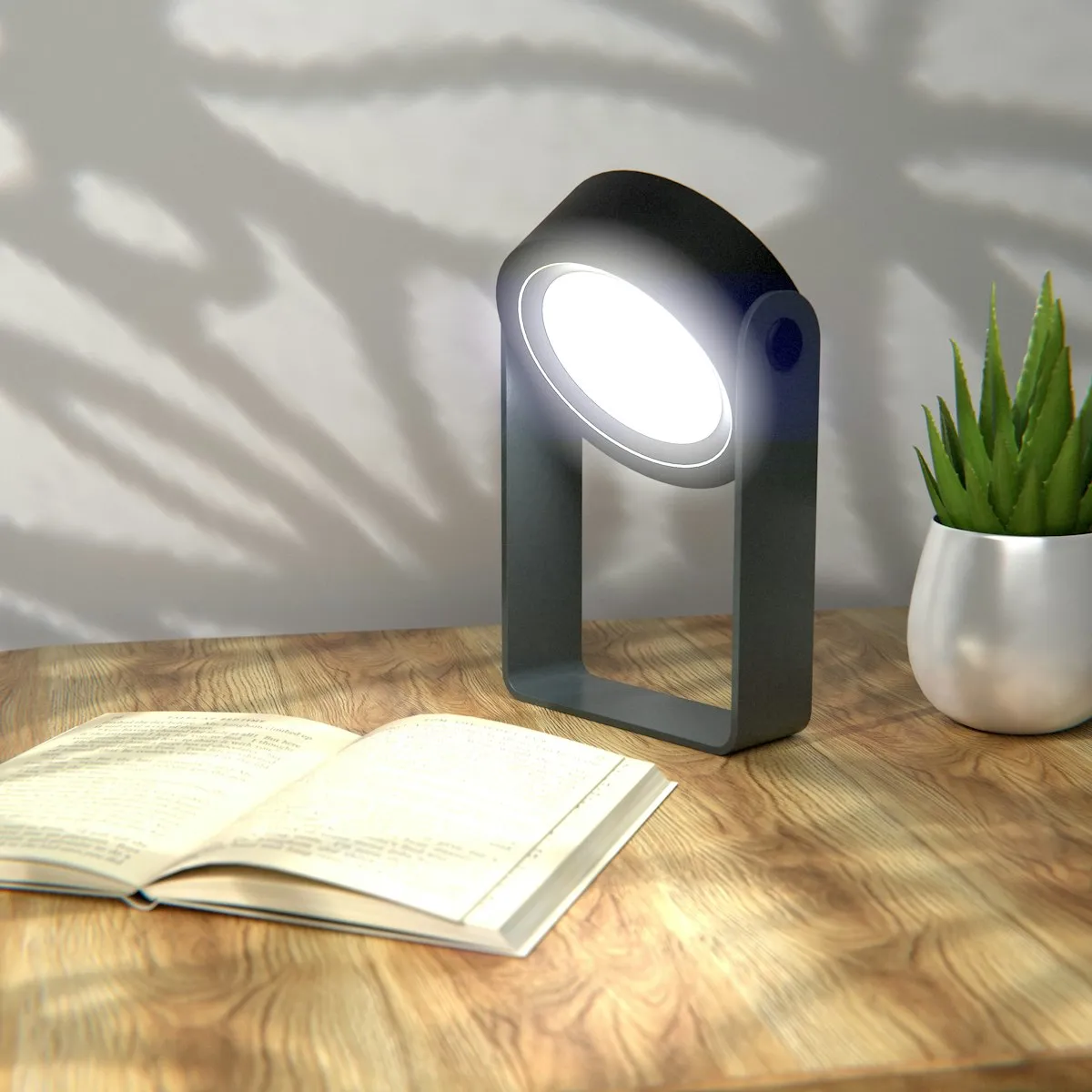 A dark grey portable LED lamp with a rounded frame and a circular light source, standing on a wooden desk beside an open book and a potted succulent.