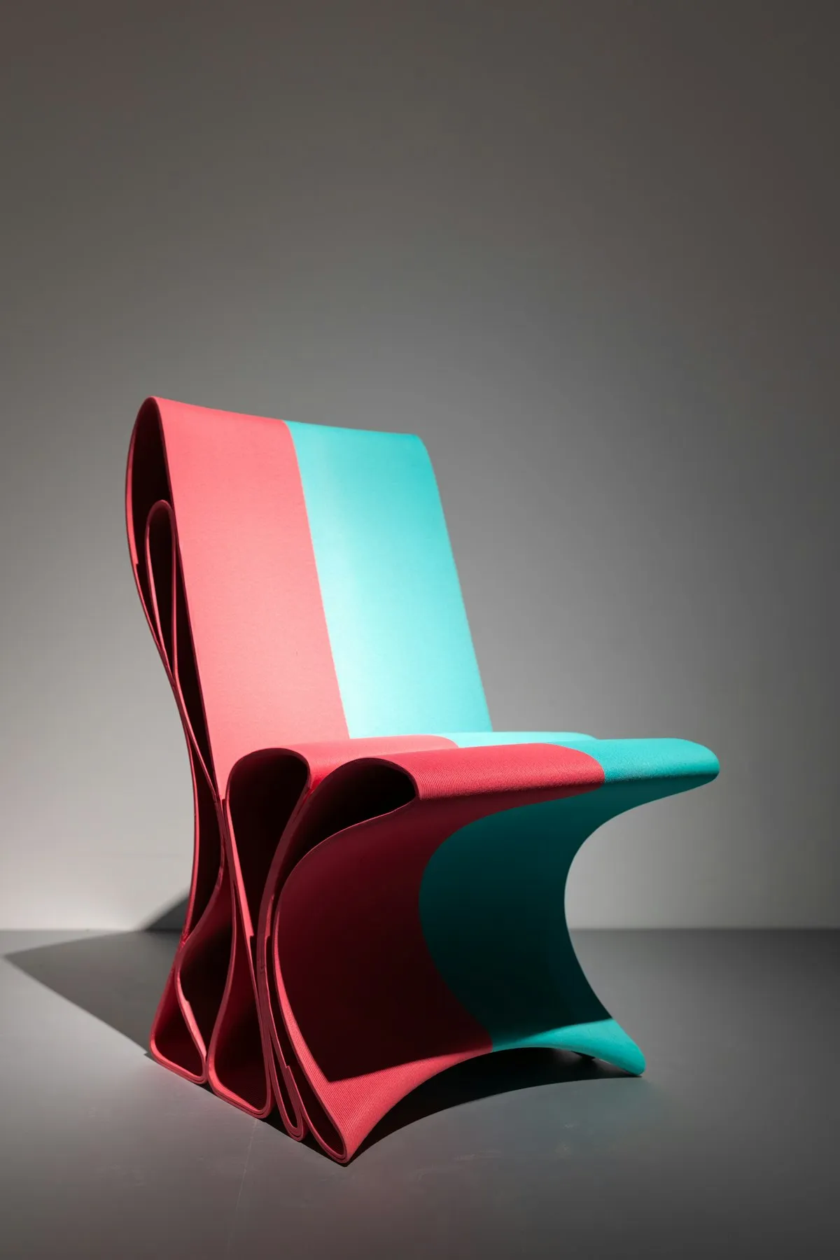 Angular chair upholstered in coral pink and turquoise fabric, made from a single pleated and folded panel that forms the backrest, seat, and legs, on a plain grey studio background.