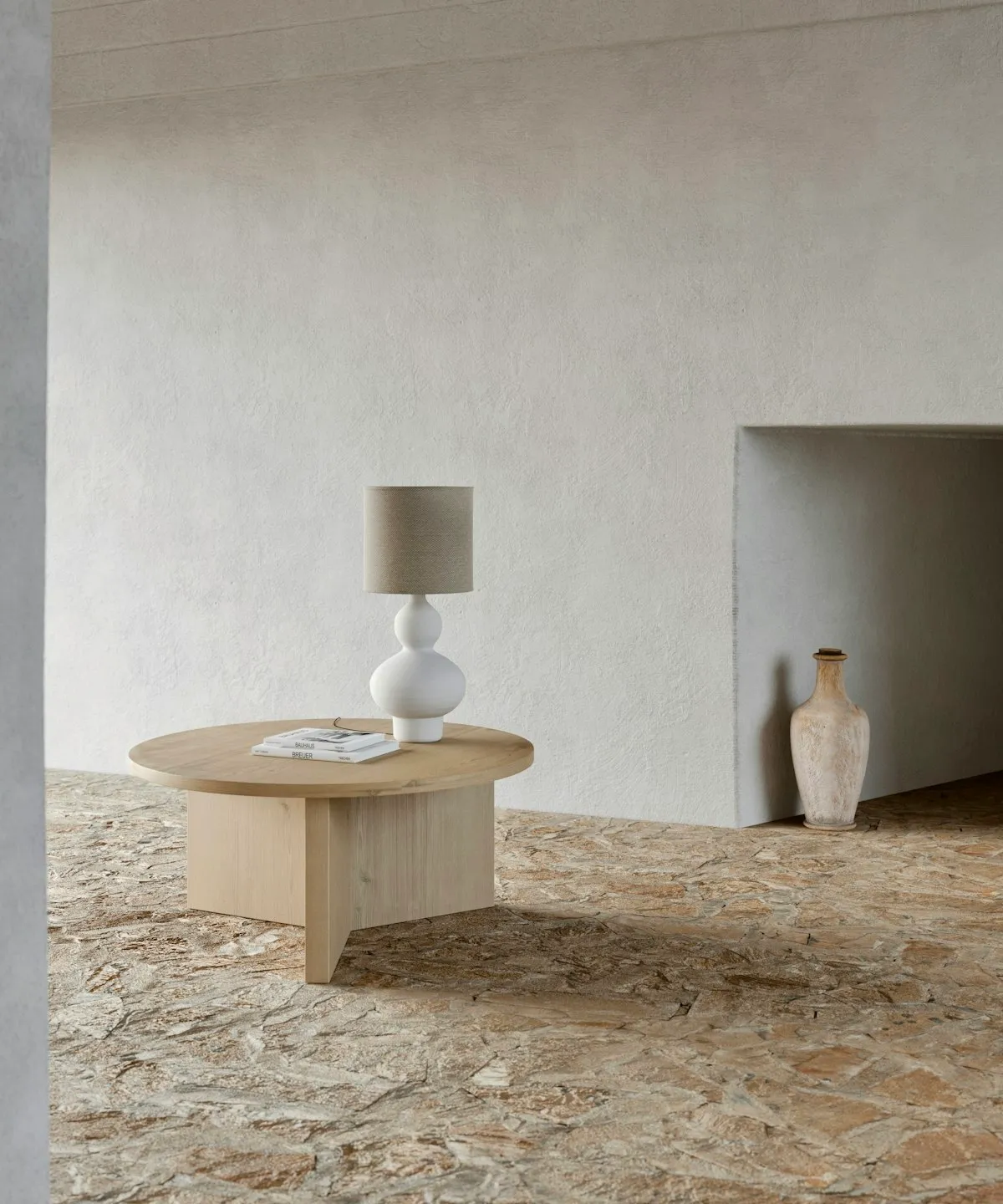 A round-bodied white ceramic table lamp with a linen drum shade, standing on a wooden coffee table in front of a plaster wall.