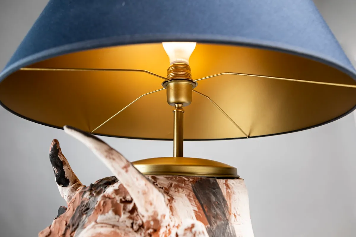 Close-up underneath a table lamp shade, showing a navy fabric exterior with a gold-lined interior, brass gallery, and driftwood base.